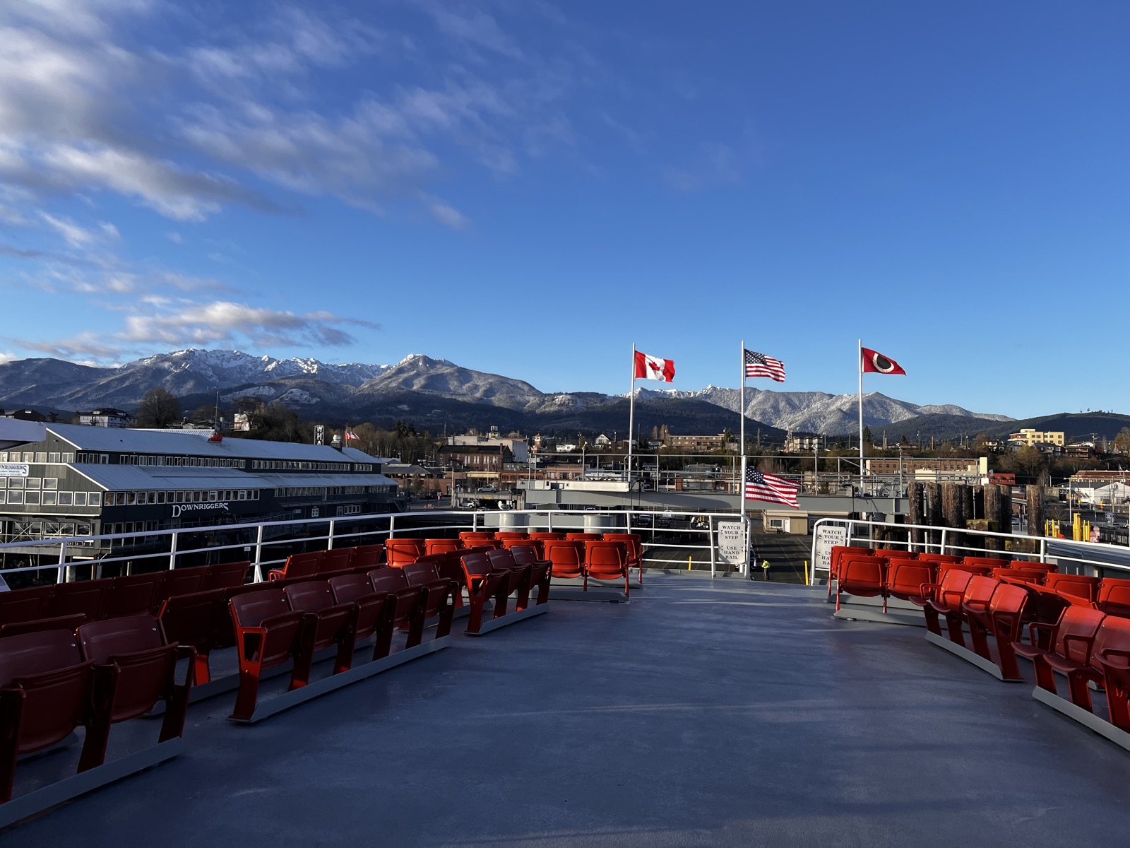 The open upper deck of a ferry with rows of empty orange seats, snow-capped mountains and a small waterfront town across the water under a blue sky